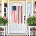 Decorative American flag hanging on a white door with plants and small tables on either side.